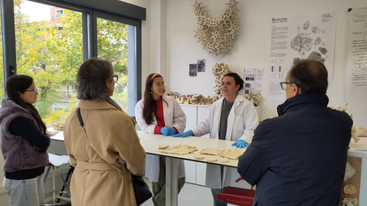 En un laboratorio luminoso, dos personas con bata blanca explican unas muestras de biomateriales sobre la mesa a un pequeño grupo de visitantes que las rodean y escuchan de pie.
