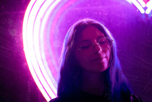 Mujer joven con expresión relajada y gafas frente a un fondo oscuro con luces rosadas de neón. Imagen de Julián Amé en Pixabay